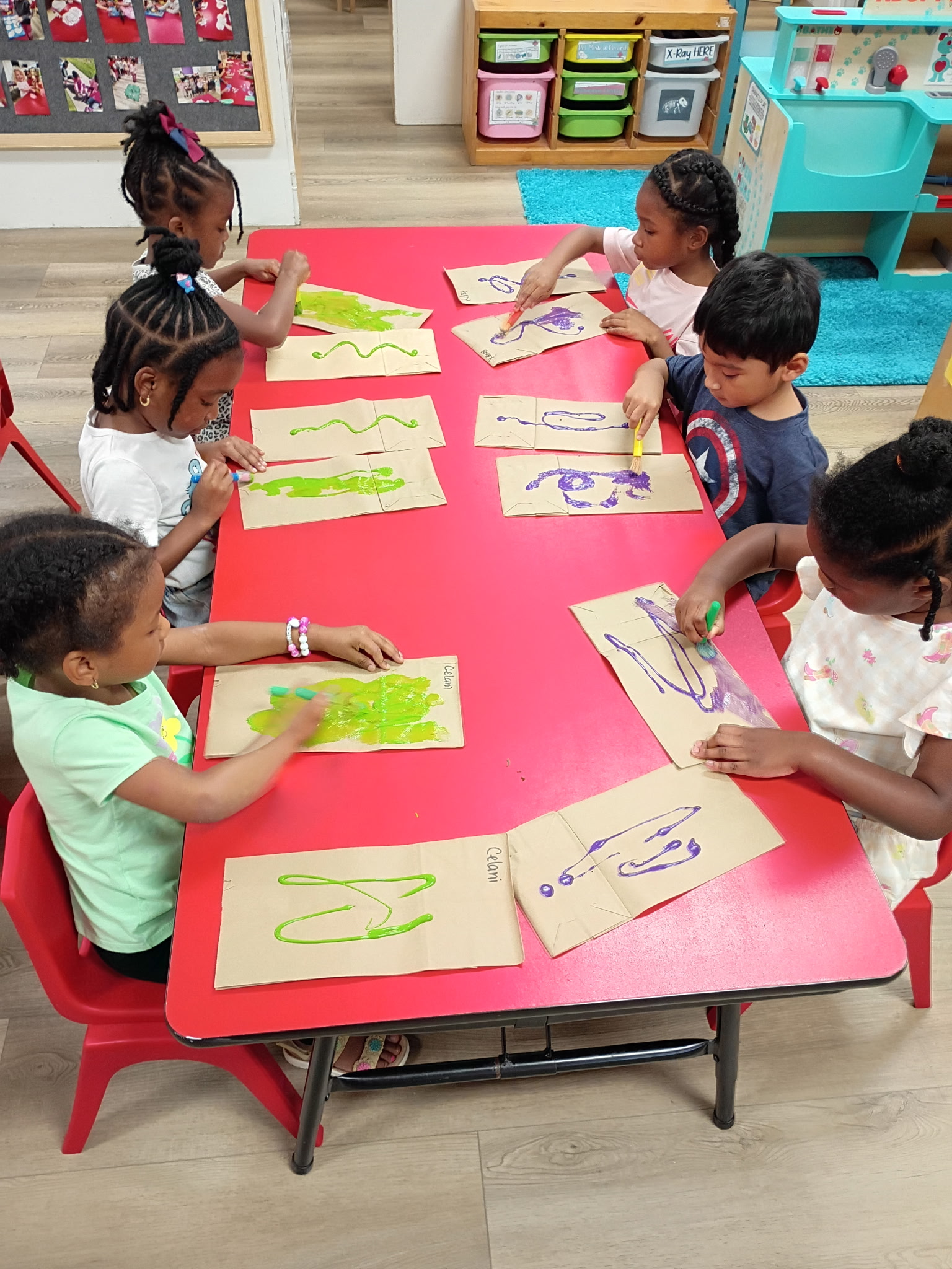 6 Children sit at a table painting brown paper bags.